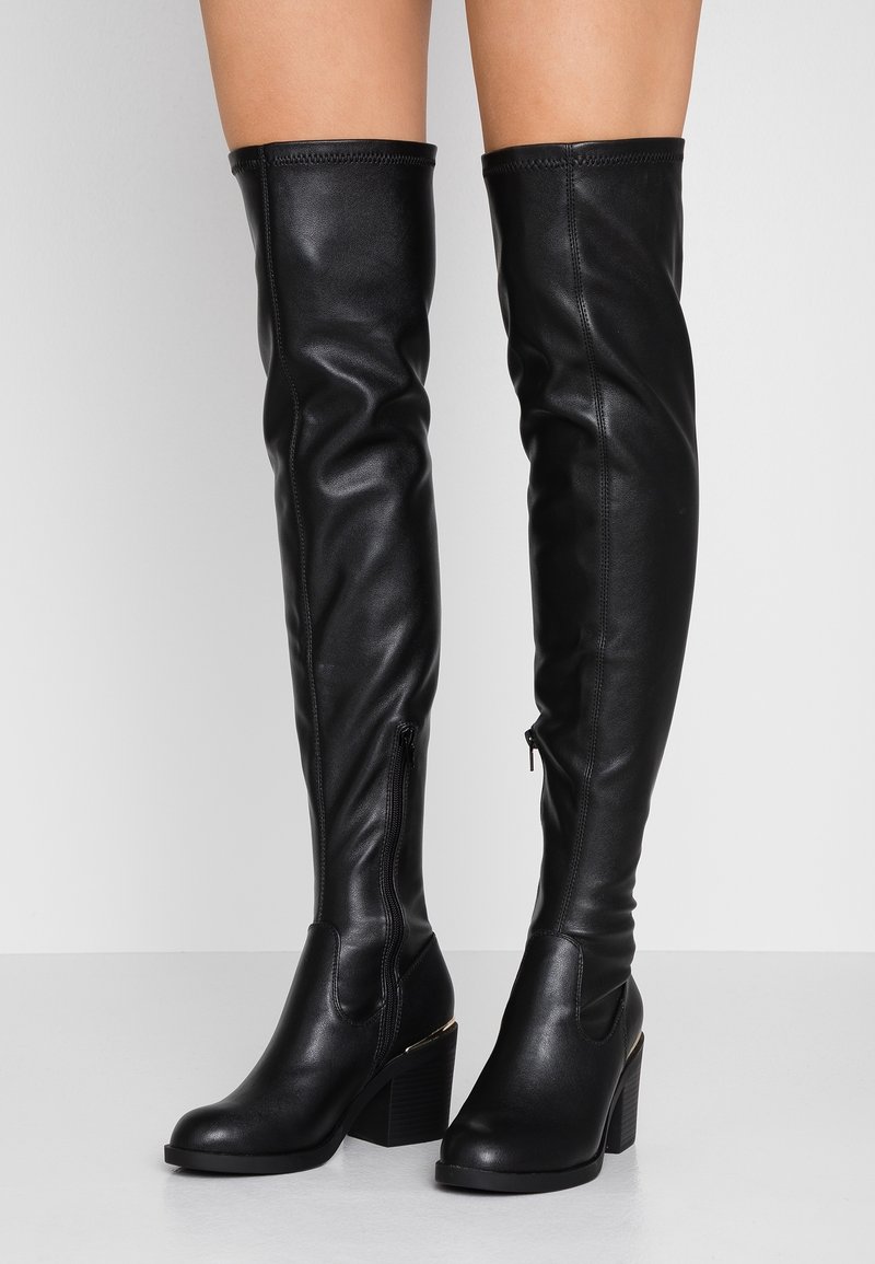 Piernas con botas de cuero negro por encima de la rodilla, con tacones en bloque y cremalleras laterales, de pie contra un fondo claro y liso.