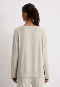 Femme avec les cheveux bruns en queue de cheval, portant un haut à manches longues beige clair et un pantalon assorti, faisant face à un mur léger et uni.