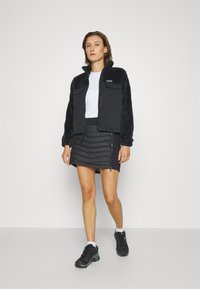 Czarna polarowa kurtka z dwiema kieszeniami na piersiach, w zestawieniu z czarną puchową spódnicą z zamkami błyskawicznymi oraz czarnymi butami sportowymi z szarymi akcentami.