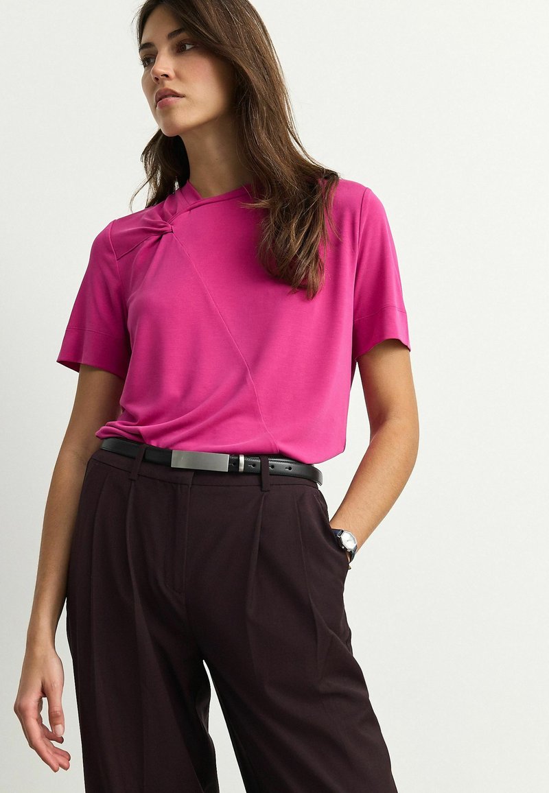 Fuchsia shirt met korte mouwen en een unieke twistdetail bij de halslijn, gecombineerd met donkere bruin hoge taillebroeken en een zwarte riem.