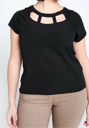 Frau trägt ein schwarzes Kurzarm-Oberteil mit Ausschnitt und beige Hose mit kleinen dunklen Mustern.