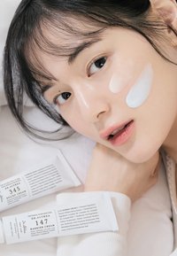 To hvite kremtuber merket "Dr. Althea 345 Relief Cream" og "Dr. Althea 147 Barrier Cream" ved siden av en person med krem på kinnet.