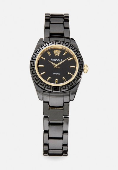 Zwarte Versace horloge met een gouden bezel en leeuwenlogo. Beschikt over een geometric patroon band, uurmarkeringen en een zwarte wijzerplaat met gouden accenten.