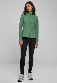 Žalia poliarinė striukė su užtrauktuku, prigludusiu dizainu, dviem šoninėmis kišenėmis ir aukštu apykakliu, derinama su juodais aptaisytais džinsais ir sportiniais sportbačiais.