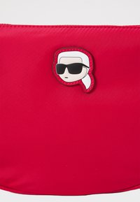 Sacca in nylon rossa con un patch cucito raffigurante un personaggio con capelli bianchi e occhiali da sole. Texture liscia con chiusura a zip.