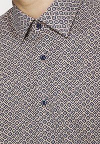 Chemise beige avec un motif géométrique de losanges bleu foncé, boutonnée avec des boutons bleu foncé, près du cou et du col d'une personne.