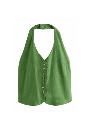 Kobieta stojąca w pomieszczeniu blisko schodów, ubrana w zielony bezrękawnik zapinany na guziki, szerokie białe spodnie i beżowe sandały, patrząca na bok.