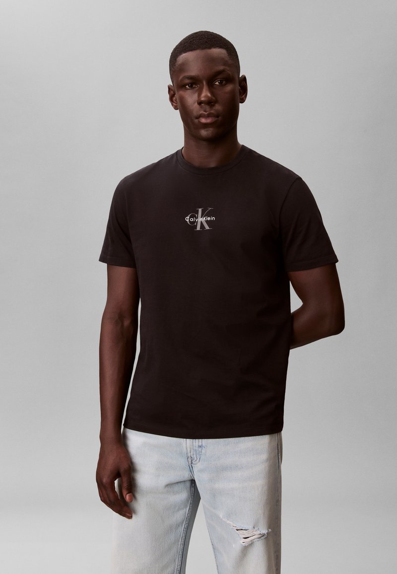 Mladý muž nosící černé tričko Calvin Klein a světle modré roztrhané džíny, stojící s neutrálním výrazem proti jednobarevnému šedému pozadí.