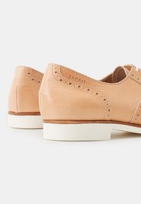 Valkoiset nahkakengät vaaleanruskeassa sävyssä, joissa on brogue-tyyliset yksityiskohdat, valkoinen pohja ja logo sivussa. Pintamateriaali on sileä ja varren muoto pyöreä.