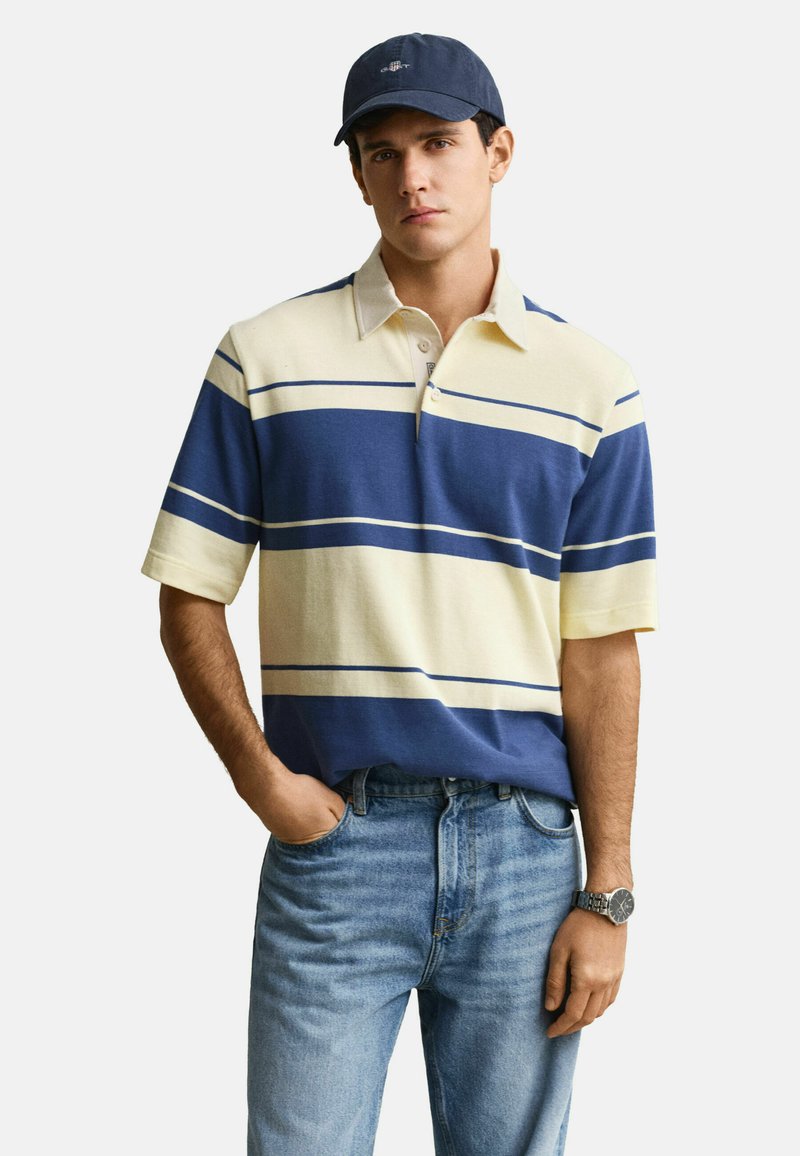 Jeune homme portant un polo à rayures bleues et crème, un jean bleu clair, une casquette marine et une montre-bracelet argentée, debout avec une main dans la poche.