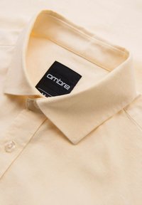 Chemise beige à col avec bouton et étiquette noire visible indiquant le nom de la marque ombre à l'intérieur du col.