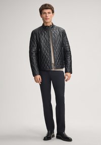 Uomo in piedi che indossa una giacca in pelle trapuntata nera sopra un maglione beige, pantaloni neri e scarpe eleganti nere su uno sfondo neutro.