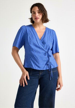 Kvinde iført en blå slå-om bluse med hvide prikker og korte ærmer, kombineret med mørkeblå jeans, stående mod en ensfarvet baggrund.