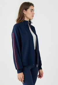 Giacca navy con zip e colletto alto, caratterizzata da dettagli a righe rosse e bianche sulle maniche. Tessuto morbido, vestibilità rilassata e polsini elasticizzati.