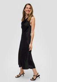 Czarna satynowa sukienka z dekoltem w kształcie drapowanej opaski i bez rękawów. Luźna sylwetka sięga połowy łydki. Połączona z czarnymi sandałami na wysokim obcasie.