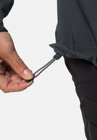 Verstelbare grijze jas met een textuurafwerking. Hand die een trekkoord met een plastic toggles vasthoudt aan de onderkant, gedragen over donkere broeken.