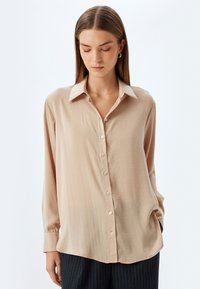 Camicia beige con bottoni realizzata in tessuto leggero, caratterizzata da colletto, maniche lunghe e una vestibilità rilassata con una leggera lucentezza e dettagli in bottoni.
