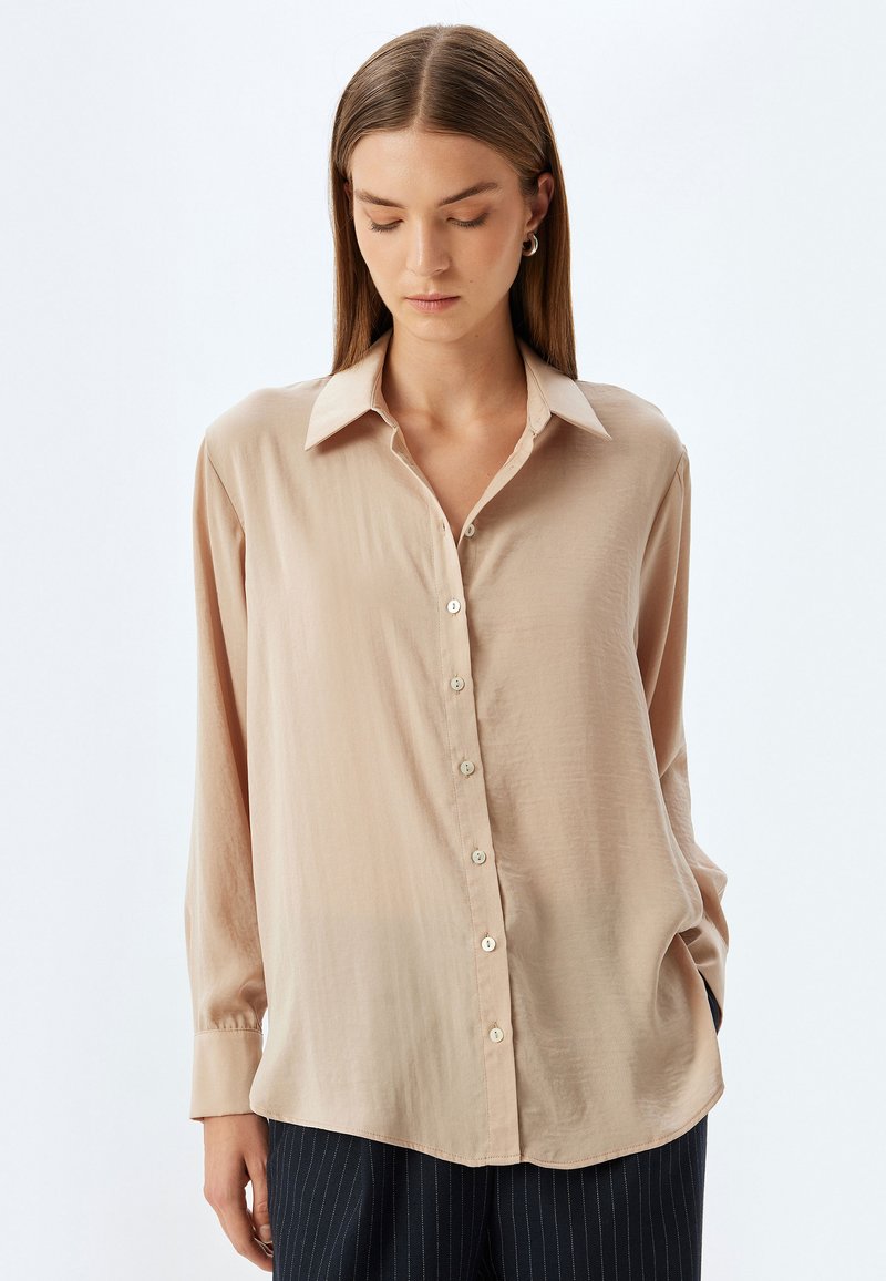 Camicia beige con bottoni realizzata in tessuto leggero, caratterizzata da colletto, maniche lunghe e una vestibilità rilassata con una leggera lucentezza e dettagli in bottoni.
