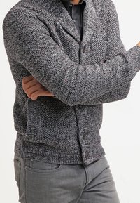 Grau gestrickter Cardigan mit strukturiertem Muster, ausgestattet mit einem Schalkragen, einer knöpfbaren Vorderseite und Taschen, kombiniert mit grauer Hose.