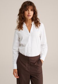 Camicia bianca con bottoni realizzata in cotone, dotata di colletto classico e maniche lunghe, abbinata a pantaloni a righe marroni. Design semplice e aderente.