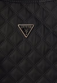 Tissu matelassé noir avec un badge métallique triangulaire portant l'inscription "GUESS U.S.A." et un point d'interrogation au centre.