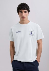 Bărbat tânăr îmbrăcat cu un tricou alb pe care este scris "Scalpers Fishing Club" și o ilustrație de pescuit pe piept, cu mâinile în buzunarele pantalonilor bleumarin.