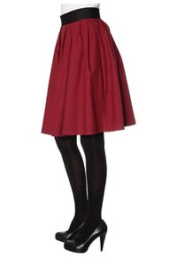 Gonna rossa al ginocchio con pieghe, cintura nera, indossata con collant neri e tacchi alti. Tessuto liscio con una forma strutturata.