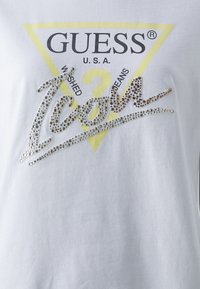 Camiseta blanca con un gráfico de triángulo amarillo que dice "GUESS U.S.A." y "WASHED JEANS." La palabra "Icon" está adornada con strass.