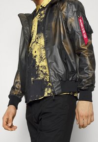 Svart bomberjacka med en glänsande yta, med huva, en dragkedjeförsedd ficka med röd etikett och ribbade muddar. Bärs över en gul mönstrad skjorta.