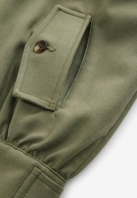Olivgrüner Stoff mit Fischgrätmuster, ausgestattet mit einer Klapptasche und einem braunen Knopf sowie einer Raffung entlang des Ärmels.