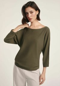 Oliwkowy, oversize'owy sweter dzianinowy z okrągłym dekoltem i rękawami do trzech czwartej; gładka tekstura i luźny krój. W połączeniu z jasnymi spodniami.
