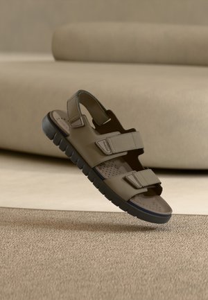 Beżowy sandał z dwoma regulowanymi paskami, gumową podeszwą i perforowaną wkładką, charakteryzujący się fakturowaną powierzchnią i minimalistycznym designem.