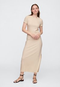 Beżowa, dopasowana maxi sukienka z krótkim rękawem, okrągłym dekoltem i bocznym rozcięciem. Noszona z czarnymi sandałami na paski. Gładka, elastyczna tkanina.