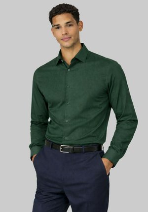 Homme portant une chemise ajustée vert foncé boutonnée, rentrée dans un pantalon bleu foncé avec une ceinture noire, les mains dans les poches, sur un fond gris neutre.