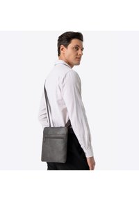 Szara skórzana listonoszka o fakturowanej powierzchni, z przednią kieszenią na suwak i regulowanym paskiem, trzymana przez osobę w jasnej koszuli.