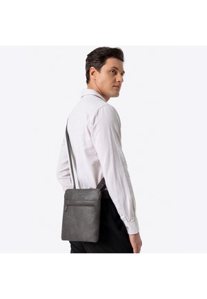 Szara skórzana listonoszka o fakturowanej powierzchni, z przednią kieszenią na suwak i regulowanym paskiem, trzymana przez osobę w jasnej koszuli.