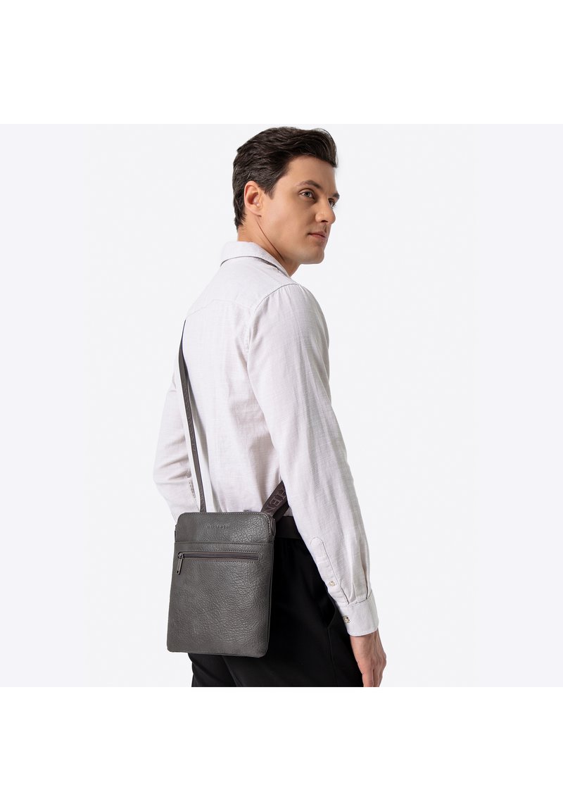 Szara skórzana listonoszka o fakturowanej powierzchni, z przednią kieszenią na suwak i regulowanym paskiem, trzymana przez osobę w jasnej koszuli.
