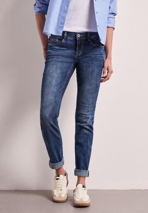 Personne portant un jean bleu retroussé, des baskets blanches, une chemise blanche et une chemise bleu clair boutonnée, debout contre un mur uni.