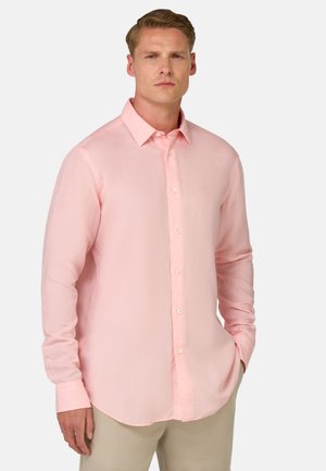 Chemise de costume à manches longues rose clair en tissu doux, dotée d'un col à boutons et d'un ourlet arrondi, portée avec un pantalon beige clair.