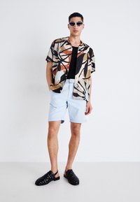 Chemise à manches courtes avec un motif abstrait coloré, associée à un short bleu clair et des chaussures noires à enfiler en tressé.