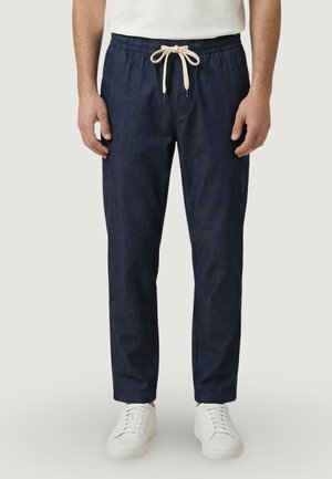 Mężczyzna w granatowych spodniach ze sznurkiem i białych butach sportowych, stojący z rękami swobodnie opuszczonymi wzdłuż ciała.