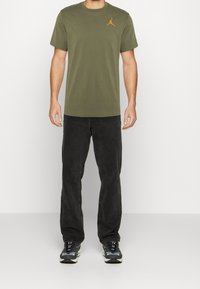 Olive green short-sleeve t-shirt with a small orange Jumpman logo on the chest, paired with black pants and black sneakers.