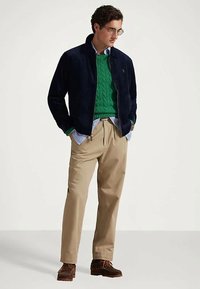 Mężczyzna w okularach, granatowej kurtce, zielonym swetrze w warkocze na niebieskiej koszuli w paski, beżowych spodniach i brązowych butach, stoi z rękami w kieszeniach.