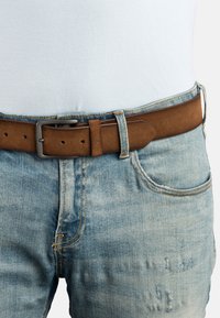 Brun struktureret bælte med en metalspænde, der bæres med blå slidte denim jeans. Nærbillede der fremhæver bæltets detaljer og pasform.