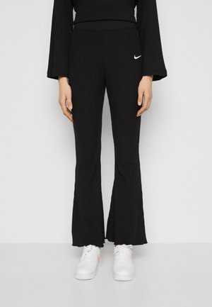 Osoba w prążkowanych czarnych spódnicach dzwonowatych z małym białym logo Nike i białymi trampkami stoi na tle jednolitego białego tła.