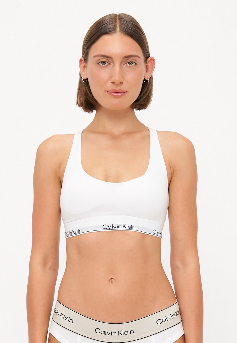 Bijela sportska grudnjak s širokim trakama i izrezom u obliku žlice, s crnim "Calvin Klein" logotipom na elastičnom pojasu. Glatki, rastezljivi materijal.