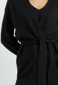 Zwarte wikkelcardigan van zacht materiaal, met een diepe V-hals, lange mouwen en een gestrikte tailleband met ribbelaccenten aan de zoom.