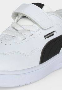 Λευκό αθλητικό παπούτσι Puma με μαύρη πλαϊνή λωρίδα, λευκά κορδόνια και αυτοκόλλητο ιμάντα σε λευκό φόντο.