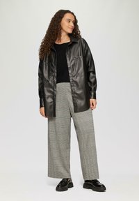 Zwarte nepleer shirtjacket, met knopen en twee borstzakken; gecombineerd met grijs geruite wijde broek en zwarte plateau schoenen.