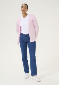 Cardigan rosa chiaro sopra una t-shirt bianca, abbinato a jeans blu e sneakers bianche. Tessuto morbido, vestibilità rilassata, silhouette allungata.